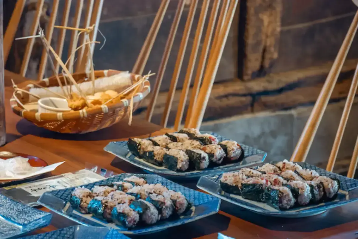Korean gimbap rolls arranged on blue ceramic plates with corn on the cob in a woven basket, displayed at an outdoor autumn dining setting with wooden chairs in the background.