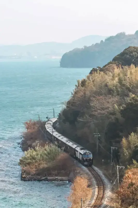 Luxury passenger train traveling along scenic coastal railway track beside turquoise waters and forested cliffs in Japan.