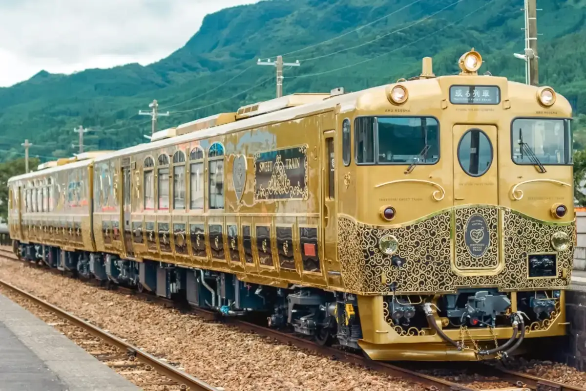 Ornate gold luxury train with decorative baroque-style patterns on its exterior, stopped at a station platform with lush green mountains in the background in Japan.