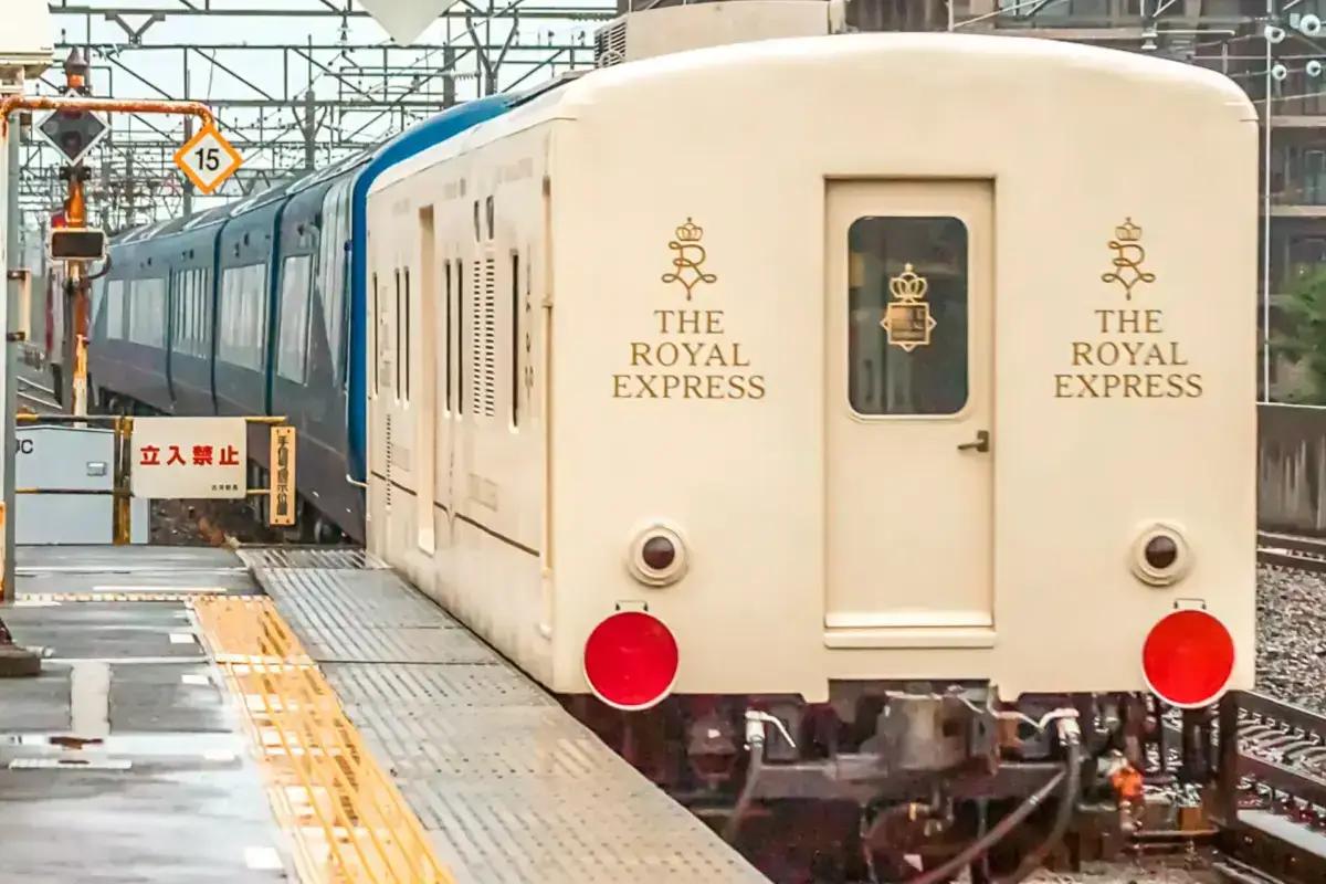 The Royal Express luxury train with cream-colored exterior and gold lettering stationed at a Japanese railway platform with overhead electrical lines.