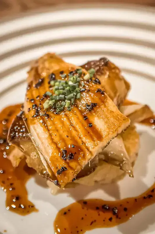 Glazed salmon fillet with caramelized golden-brown skin, garnished with sesame seeds and chopped scallions, served on a white plate with savory sauce.