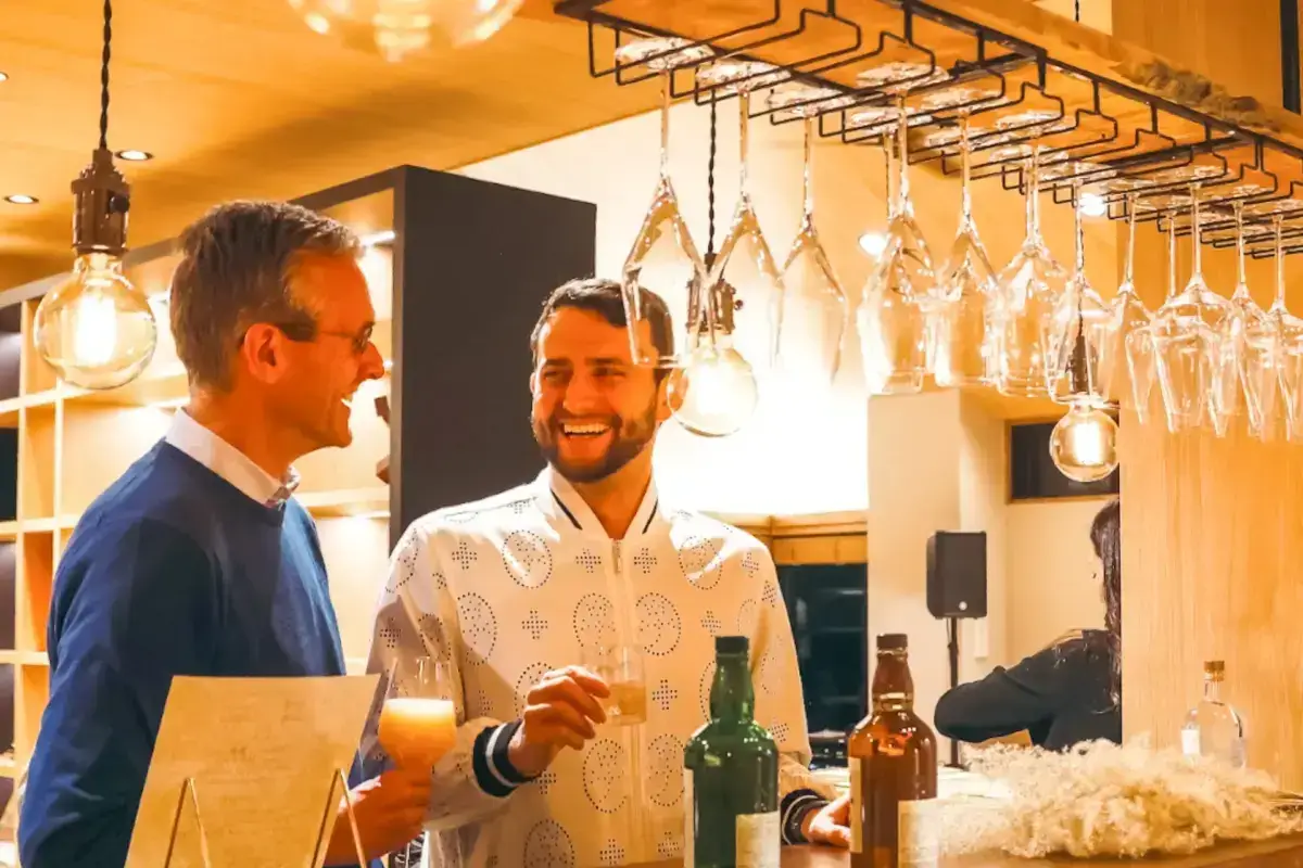 Two business professionals enjoying drinks and conversation at a stylish bar with hanging wine glasses and warm ambient lighting, fostering informal networking and relationship building.