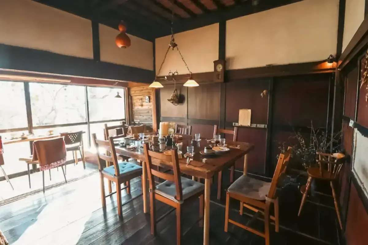 Warm rustic dining room with wooden table set for a meal, featuring exposed beam ceiling, dual pendant lighting, large windows with natural light, and vintage decor accents.