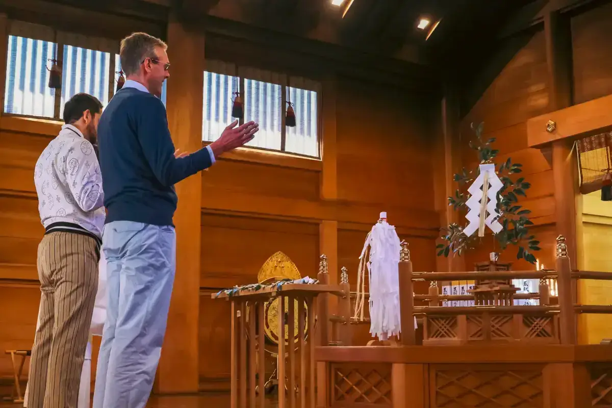 Two men observing a traditional Japanese Shinto shrine altar with sacred ornaments, paper streamers, and decorative branches in a wooden interior space