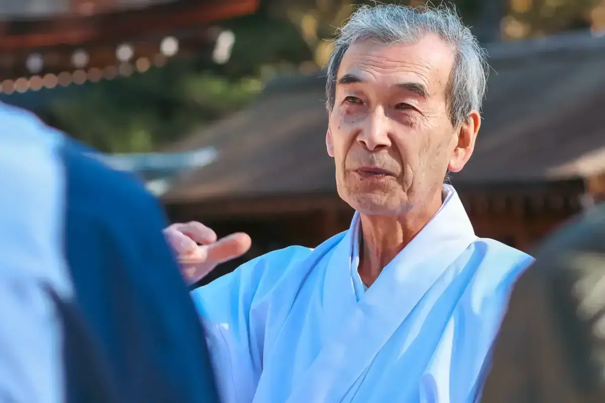 Senior Japanese man in light blue kimono engaged in conversation outdoors, gesturing with his hand in a professional or cultural setting.