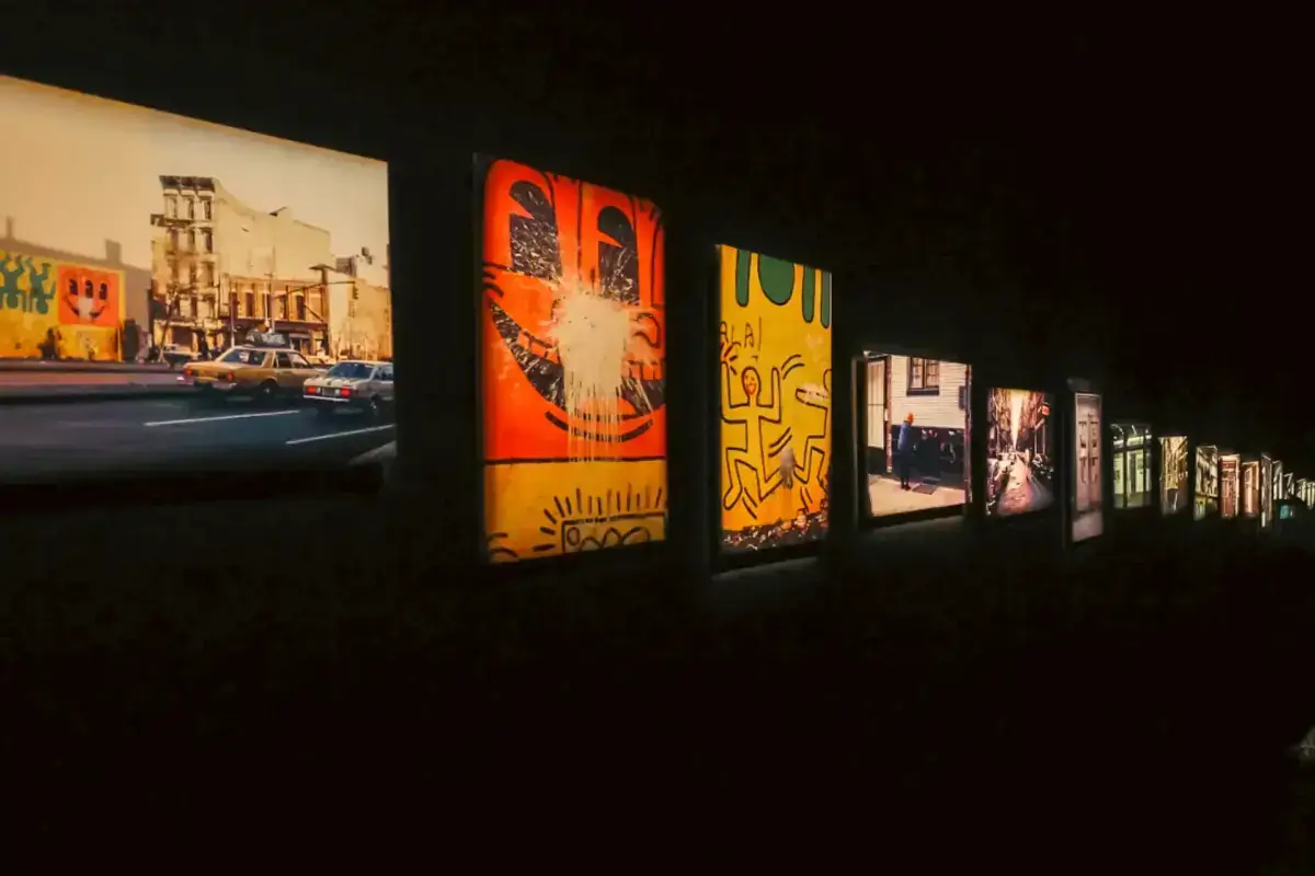Row of framed photographs and Keith Haring-style artworks displayed on a dark wall, featuring urban street scenes and colorful pop art imagery in orange and yellow tones.