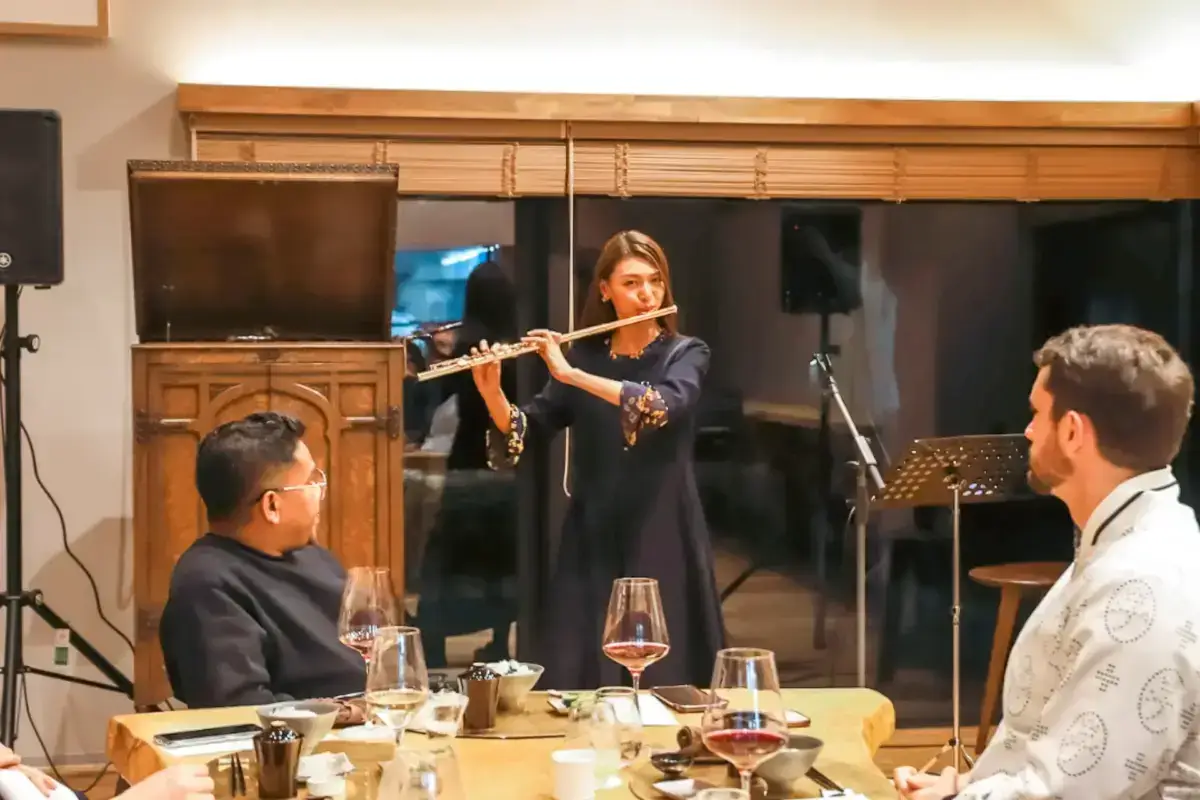 Female flutist performing for an intimate dining audience in a modern Japanese-inspired room with traditional wooden fixtures and bamboo blinds.