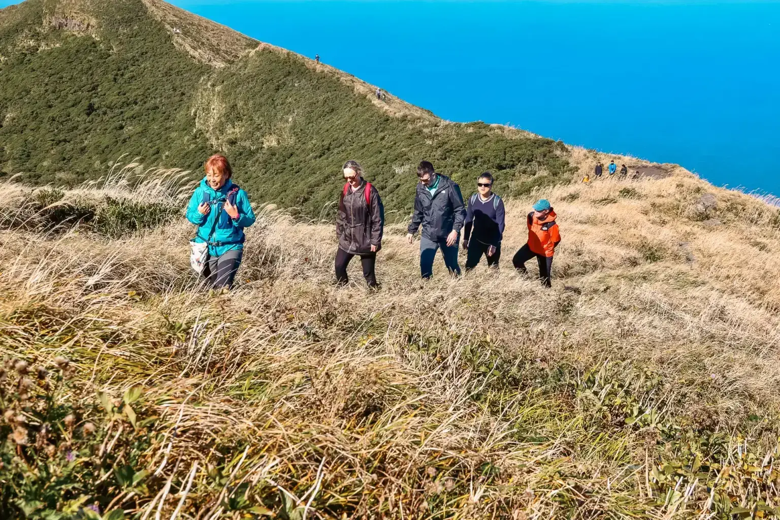 Group of hikers trekking along a grassy hillside trail on Hachijojima Island with volcanic mountains and clear blue sky in the background.