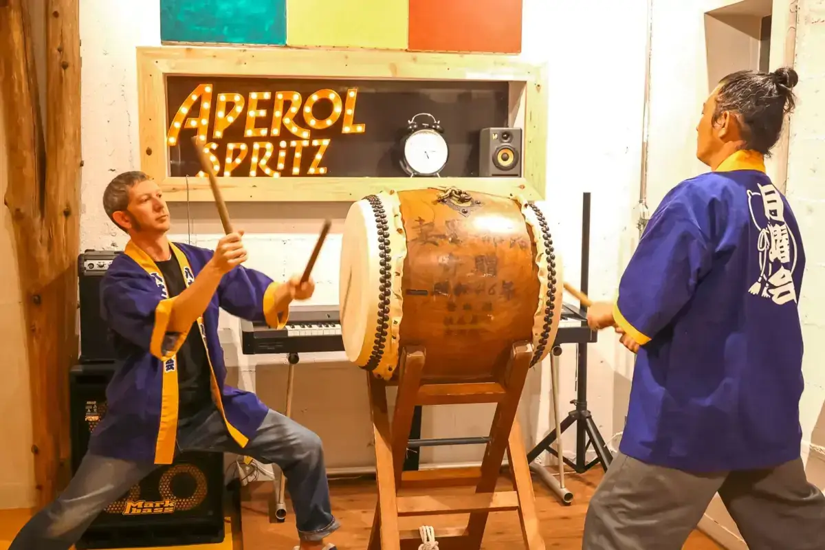Two performers in traditional blue happi coats playing a large taiko drum in a vibrant studio space, with an illuminated "Aperol Spritz" sign and colorful artwork in the background.