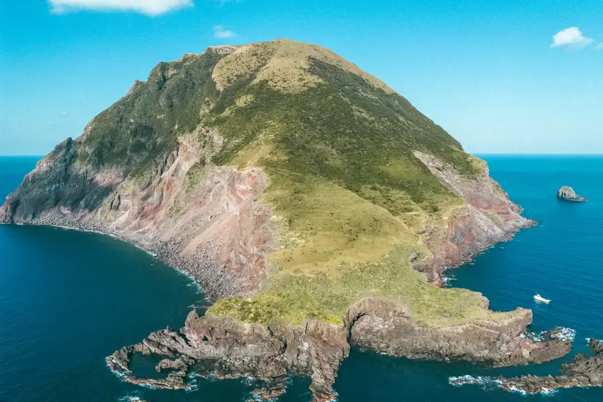 Aerial view of Hachijojima's volcanic peninsula with lush green slopes and dramatic exposed rock formations jutting into the deep blue Pacific Ocean under a clear sky.