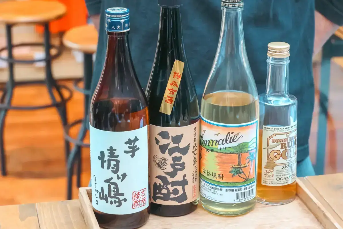 Four bottles of Japanese sake and shochu arranged on a wooden tray, featuring local Hachijojima spirits including traditional sake bottles with Japanese calligraphy labels and tropical-themed shochu bottles.