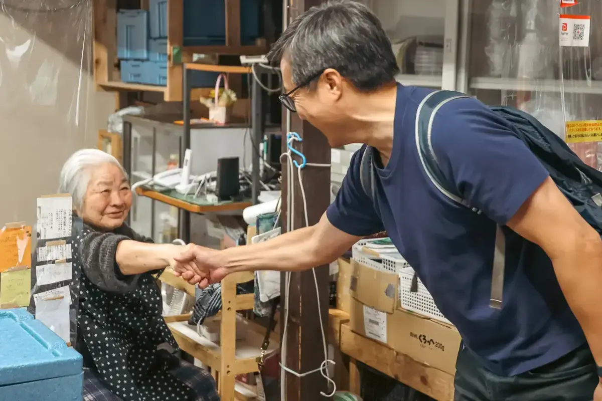 Elderly Japanese woman in traditional clothing warmly shaking hands with a visitor in a local workshop filled with crafts and supplies on Hachijojima Island.