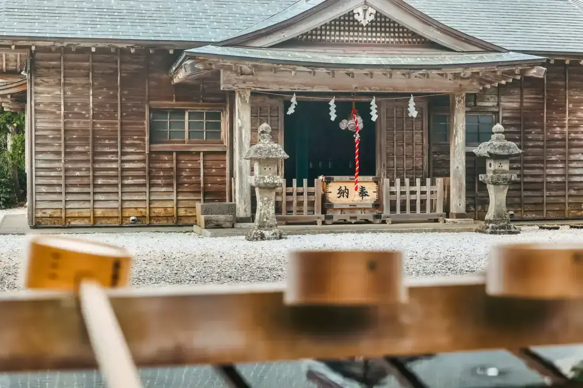 Traditional Japanese wooden shrine with stone lanterns and ceremonial decorations, viewed from a dining table with sake cups and wooden plates in the foreground at Hachijojima.