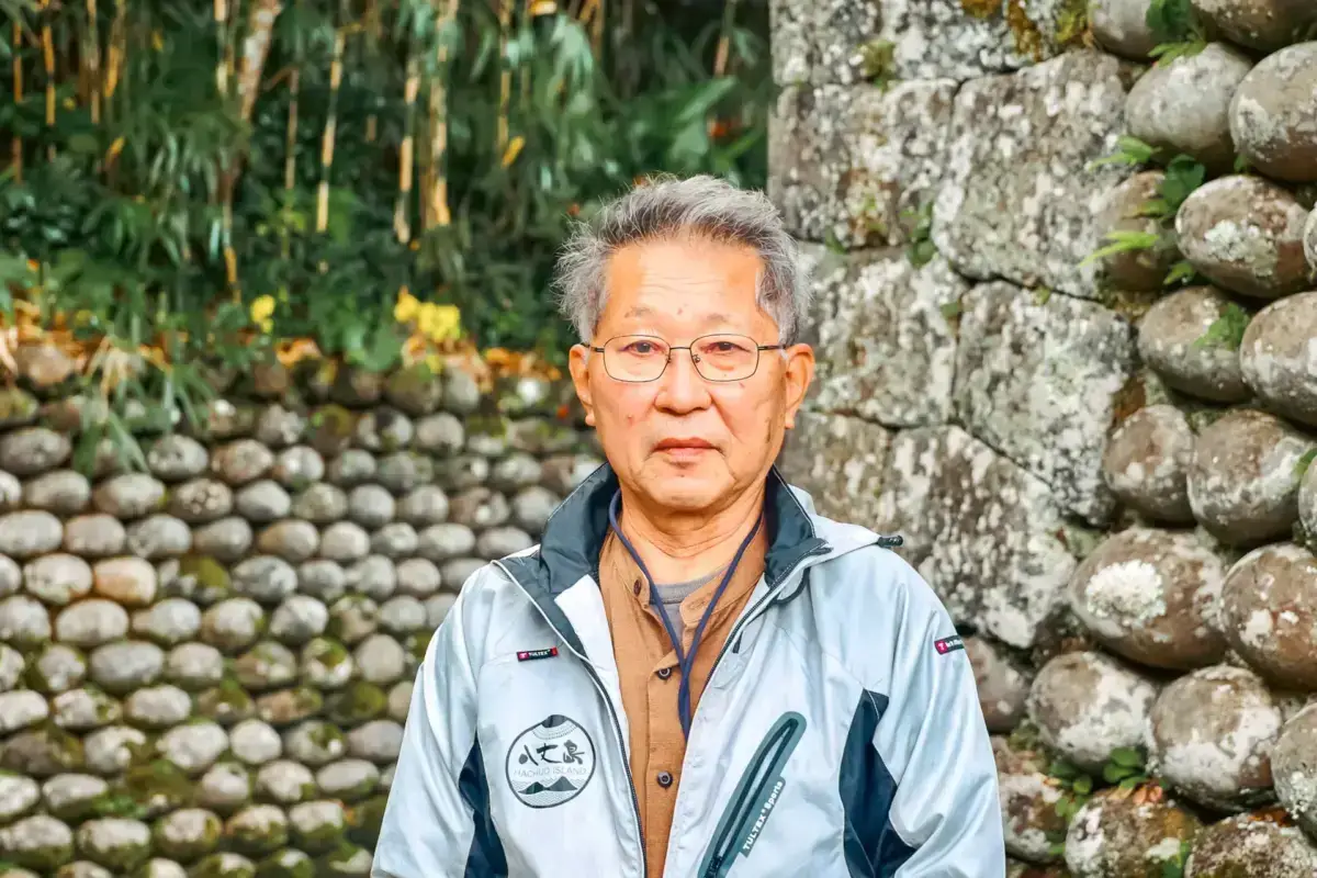Elderly Japanese man wearing glasses and a light blue jacket standing in front of traditional stone walls on Hachijojima Island.