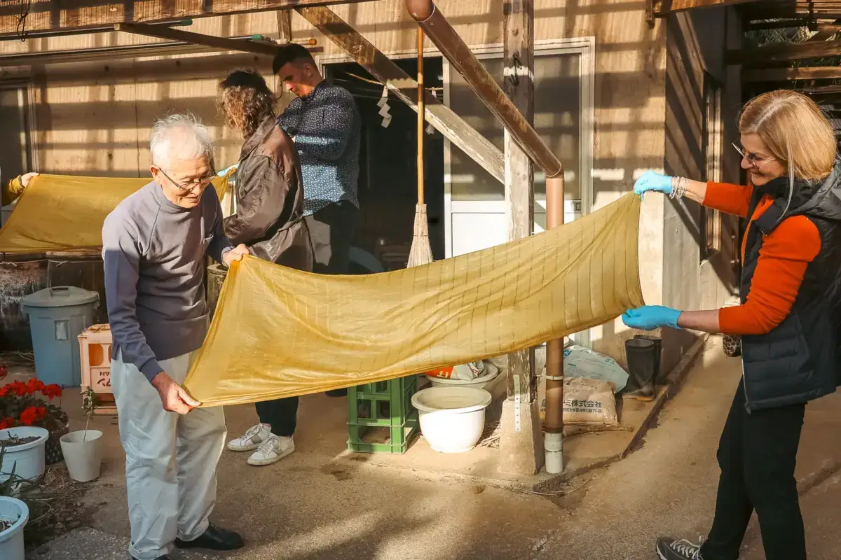 Visitors participating in traditional silk fabric weaving workshop at Hachijojima, stretching golden-yellow kihachijo silk textile in a sunlit traditional Japanese workspace.