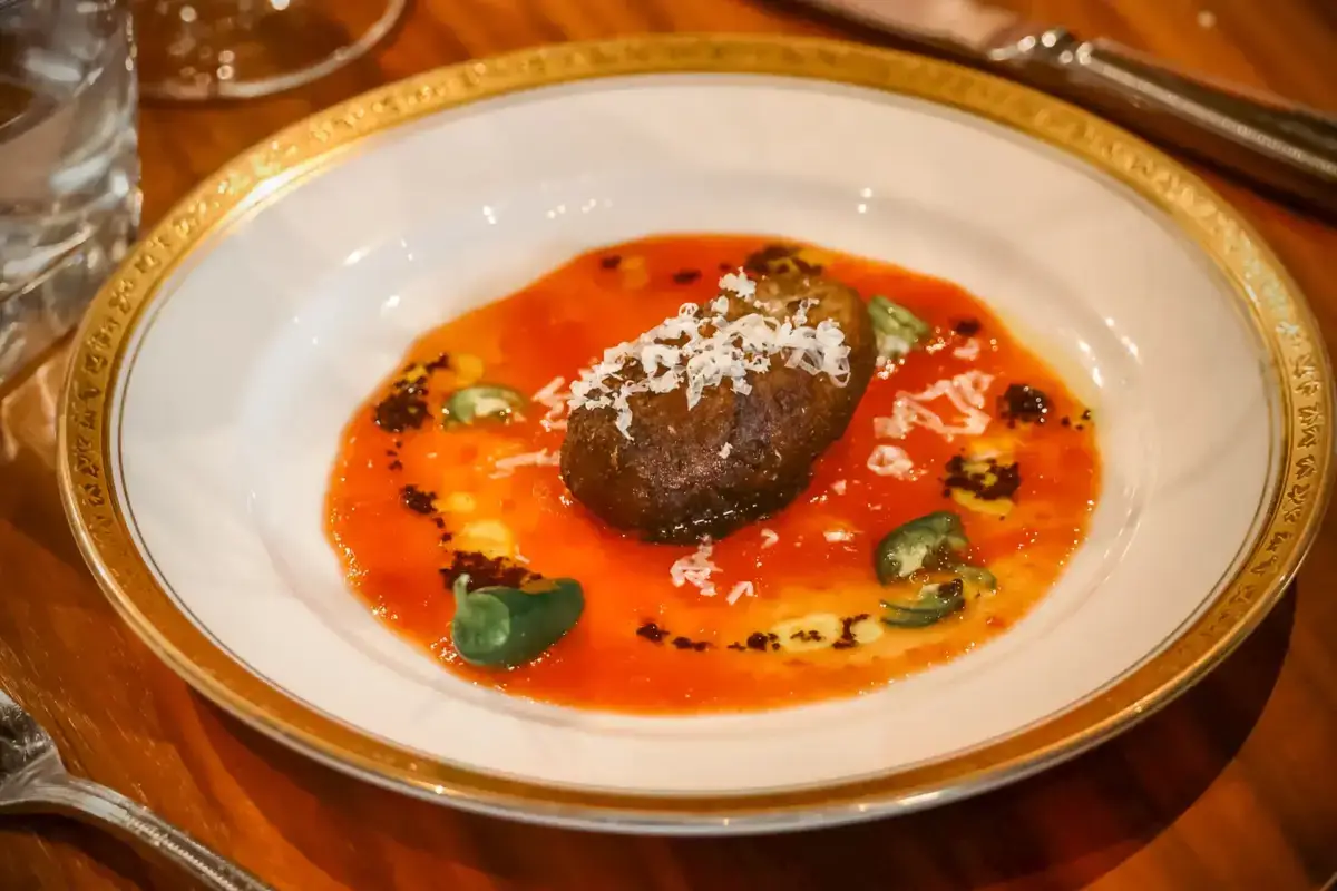 Gourmet meat dish topped with shaved cheese on a vibrant red pepper sauce, garnished with microgreens and served on an elegant gold-rimmed plate.