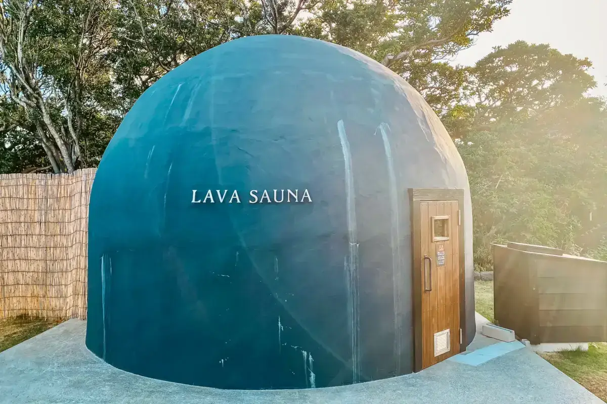 Modern blue geodesic dome-shaped lava sauna with wooden door, set against a backdrop of lush green trees in a tranquil outdoor wellness area at Hachijojima.