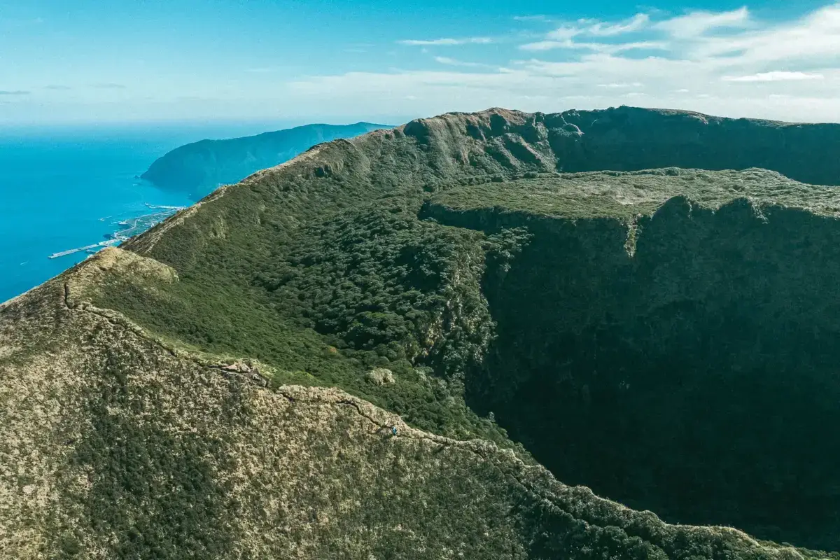 Aerial view of Hachijojima's dramatic volcanic crater rim covered in lush green vegetation, with steep cliffs descending to the turquoise Pacific Ocean under partly cloudy skies.