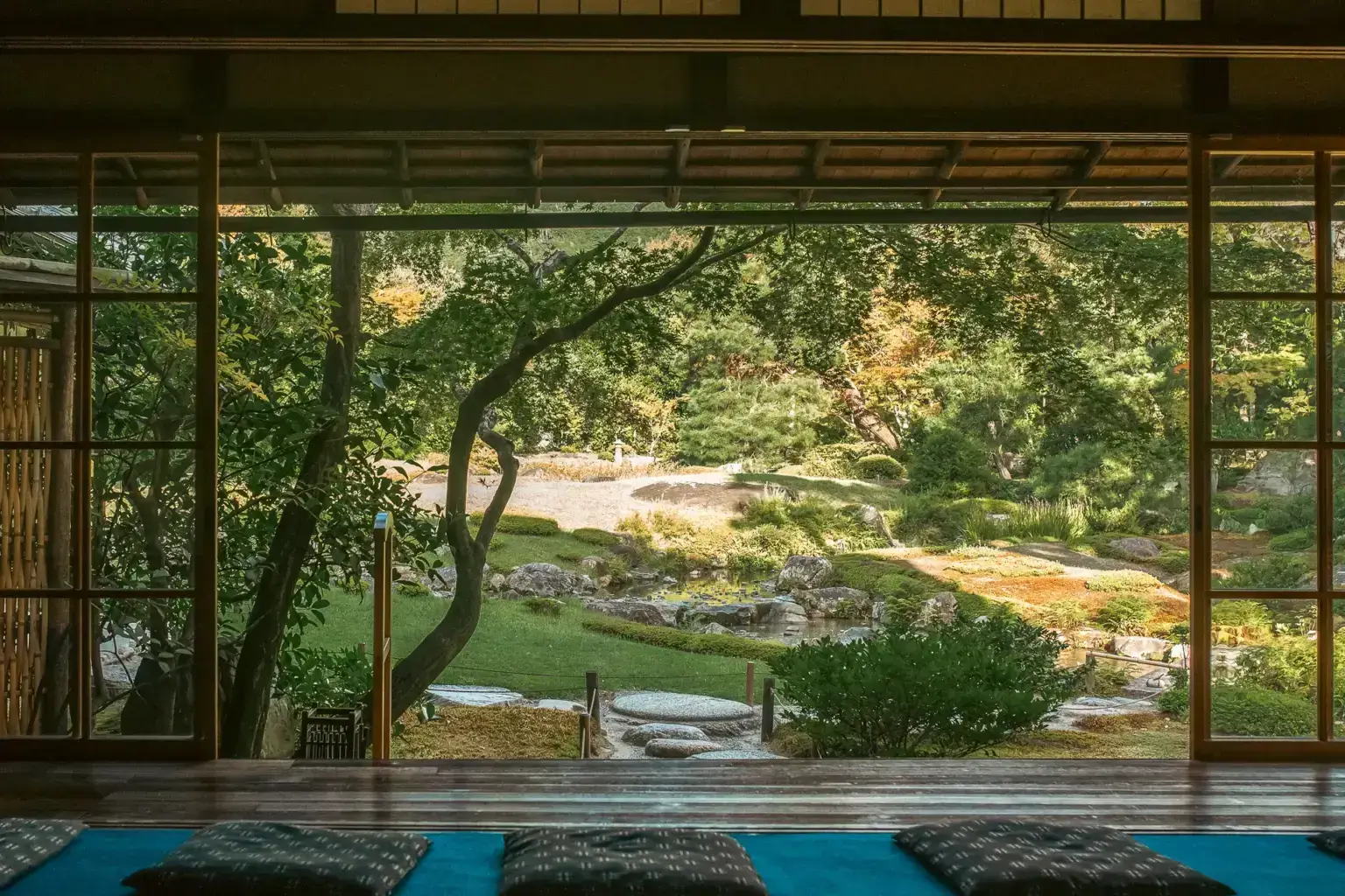 Traditional Japanese room with tatami flooring and sliding shoji doors opening to a serene garden with trees, grass, and a winding path bathed in warm sunlight.