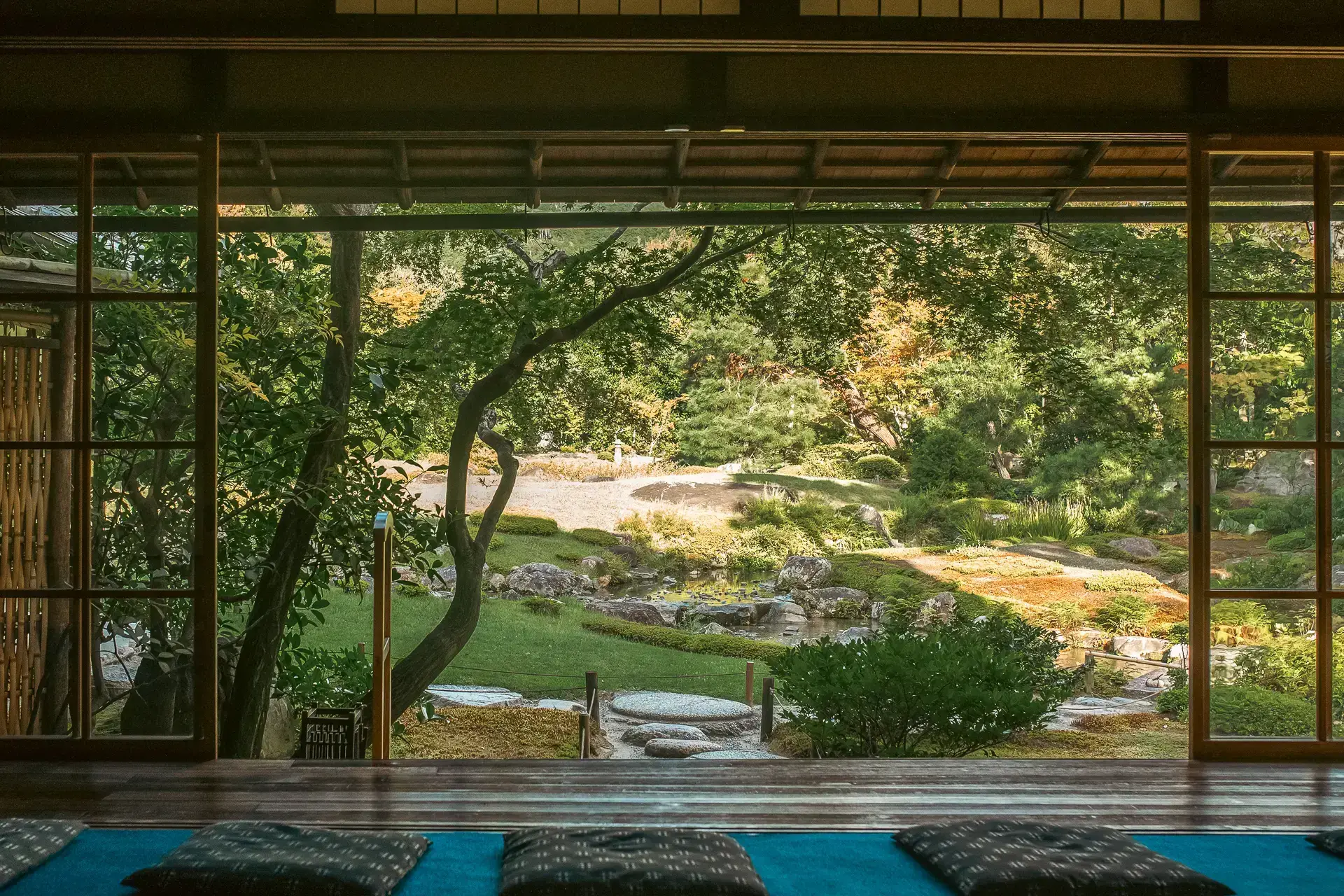 Traditional Japanese room with tatami flooring and sliding shoji doors opening to a serene garden with trees, grass, and a winding path bathed in warm sunlight.