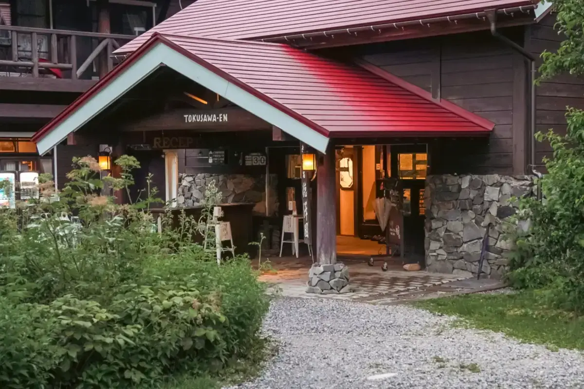 Traditional Japanese mountain lodge with stone foundation, dark wood exterior, and red roof, surrounded by lush greenery with warm interior lighting creating a welcoming atmosphere.