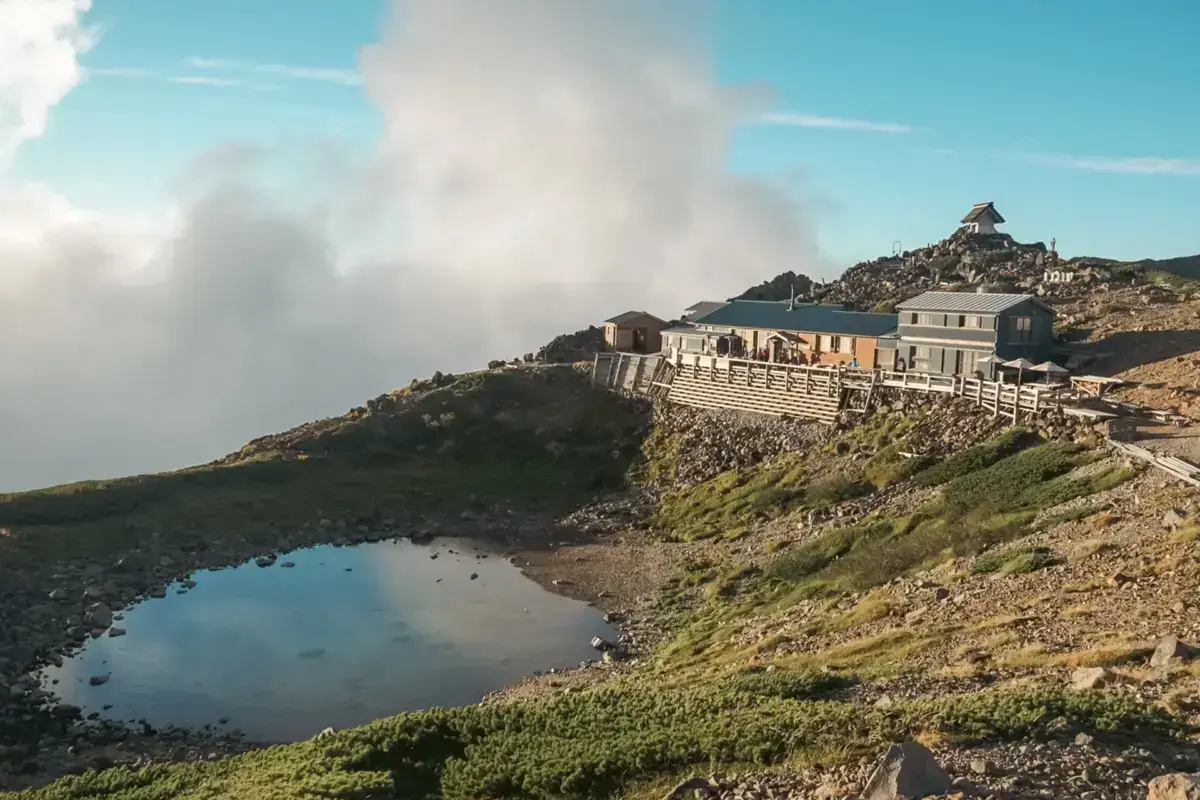 Mountain lodge buildings perched on a rocky ridge above clouds with a small blue pond in the foreground, surrounded by alpine vegetation under a clear blue sky.
