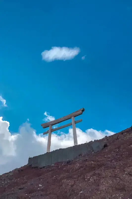 Wooden torii gate standing on volcanic mountain slope against clear blue sky with scattered clouds.