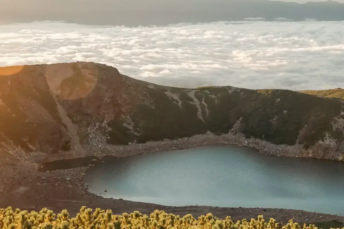 Turquoise crater lake nestled in volcanic mountain peaks, with a dramatic sea of clouds stretching across the horizon and golden sunlight illuminating the rocky terrain.
