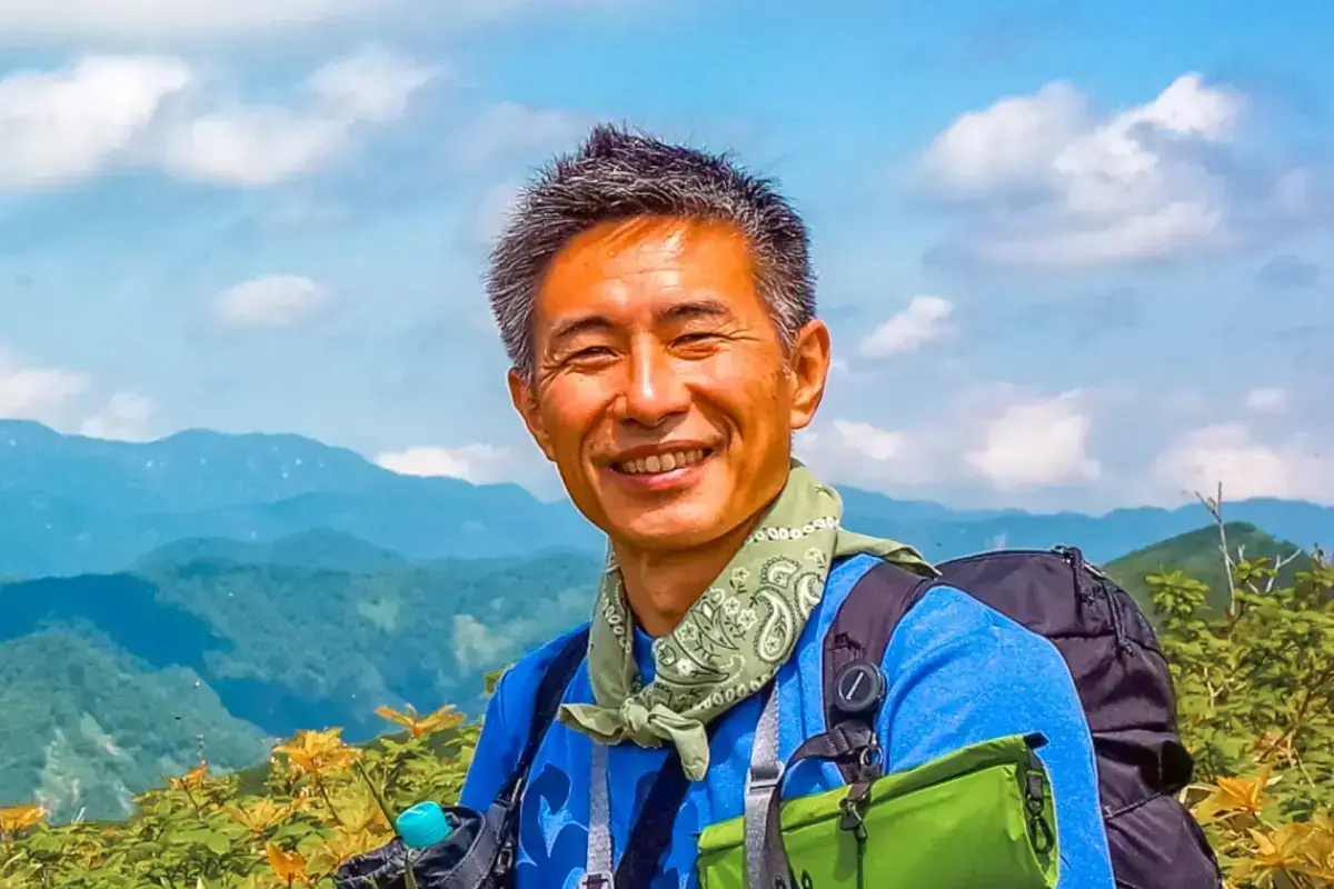 Smiling hiker with backpack and bandana posing at a mountain summit with layered blue mountain ranges and cloudy sky in the background.