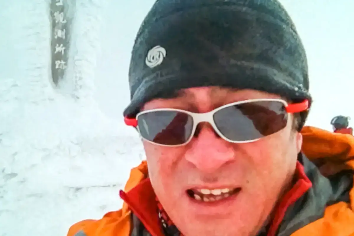 Smiling mountain climber wearing sports sunglasses and a black beanie in snowy conditions with a summit marker sign visible in the background.