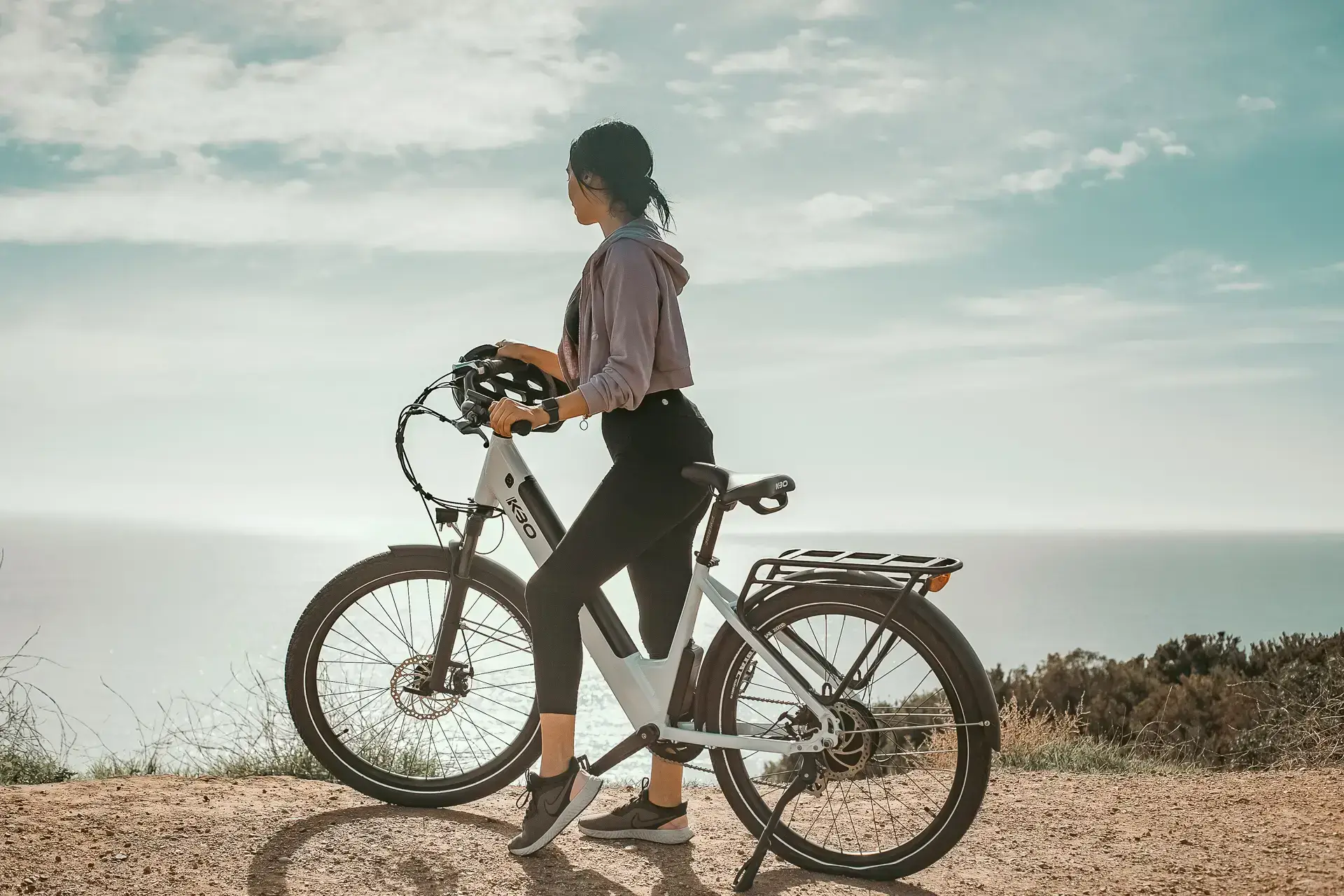 Woman with bicycle helmet standing beside white e-bike on coastal path overlooking ocean with scenic cloudy sky in Wakayama, Japan.
