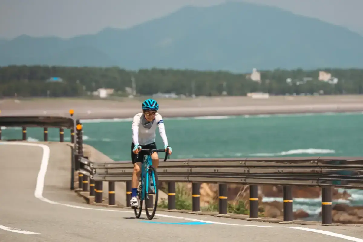 Cyclist riding a turquoise road bike along a coastal road in Wakayama, with turquoise ocean waters, sandy beach, and misty mountains in the background.