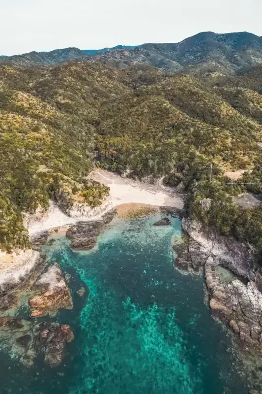 Aerial view of a secluded coastal cove in Wakayama with crystal-clear turquoise waters, white sandy beach, and rocky cliffs surrounded by lush green mountains.