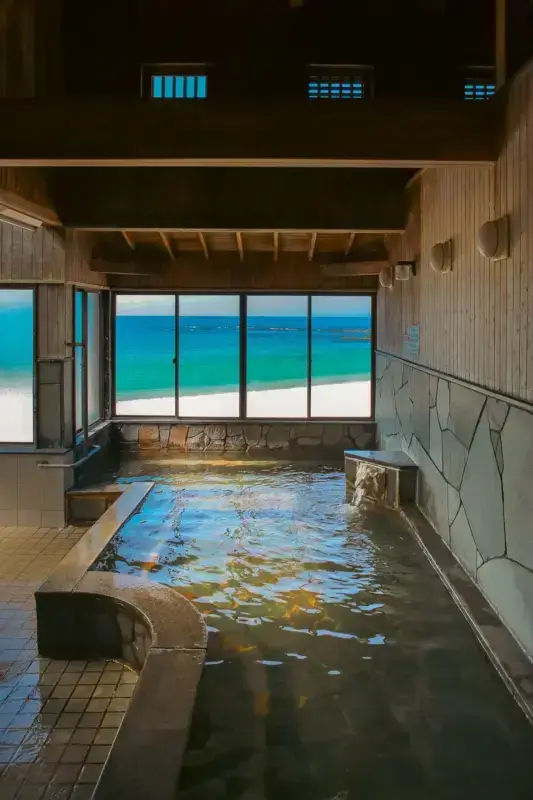 Indoor hot spring bath with stone walls and wooden ceiling overlooking turquoise ocean and white sandy beach through large glass windows in Wakayama, Japan.