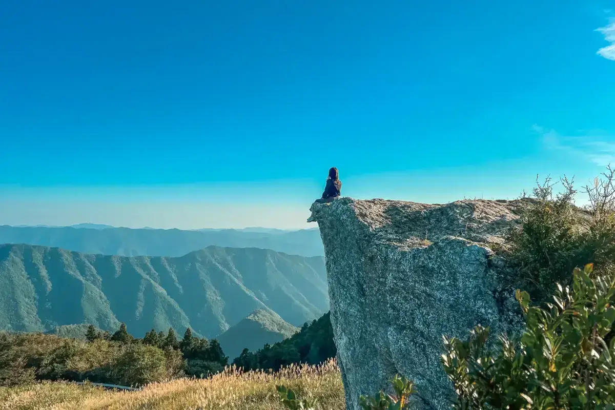 Solo hiker sitting on dramatic cliff edge overlooking layered mountain ranges in Wakayama, Japan under a clear blue sky.