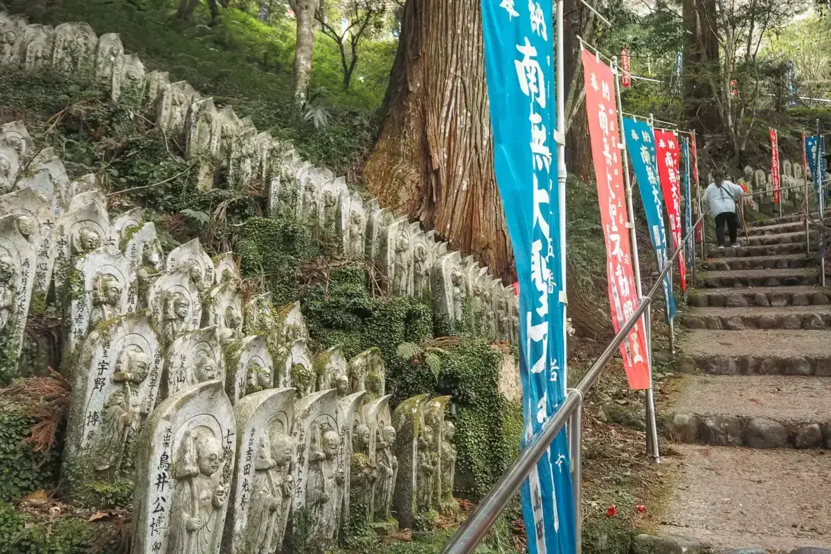 Rows of weathered stone jizo statues line a forest path leading to stone steps adorned with colorful Japanese banners at a temple site.