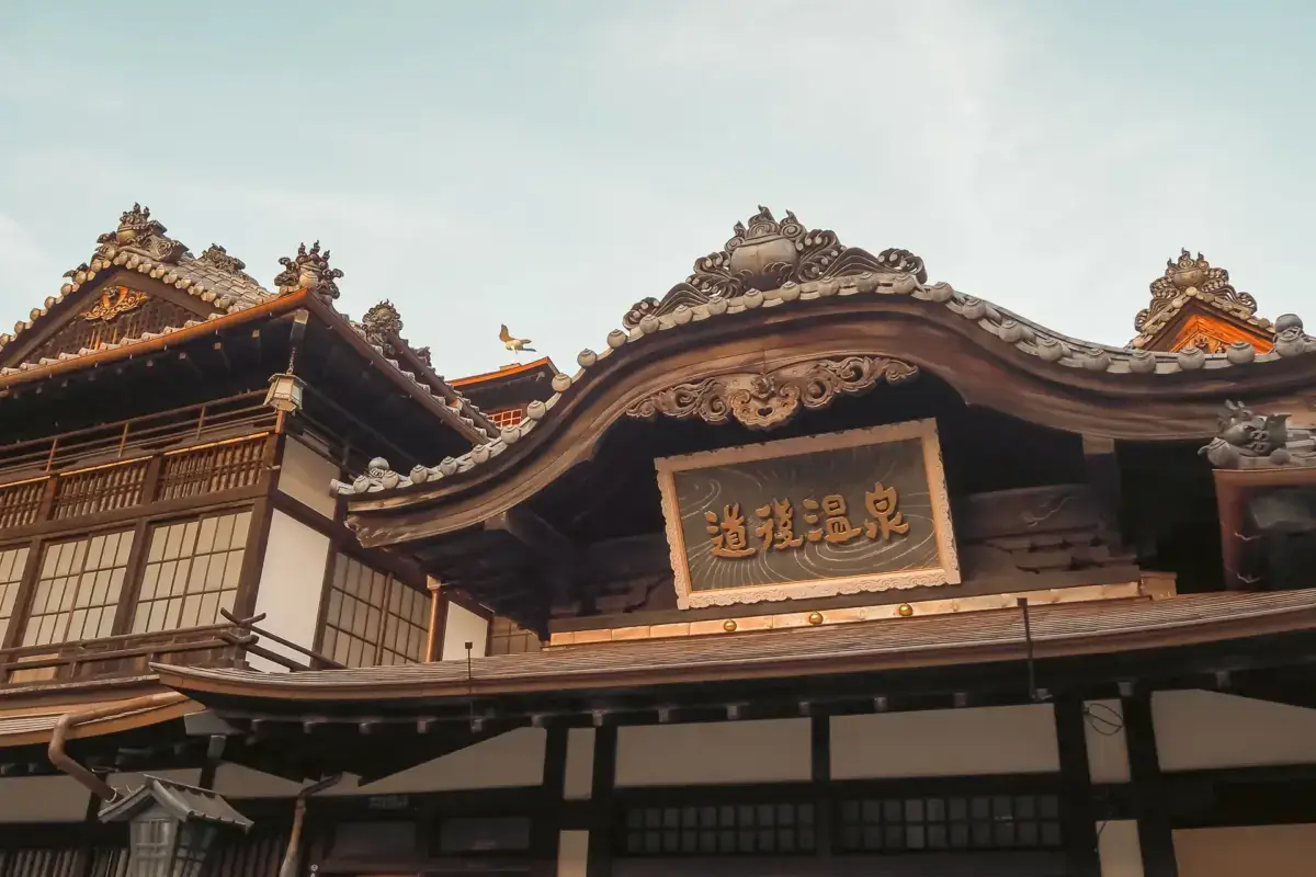 Traditional Japanese hot spring bathhouse featuring ornate curved rooflines with decorative tile eaves and a central wooden sign displaying Chinese characters, set against a tranquil evening sky.
