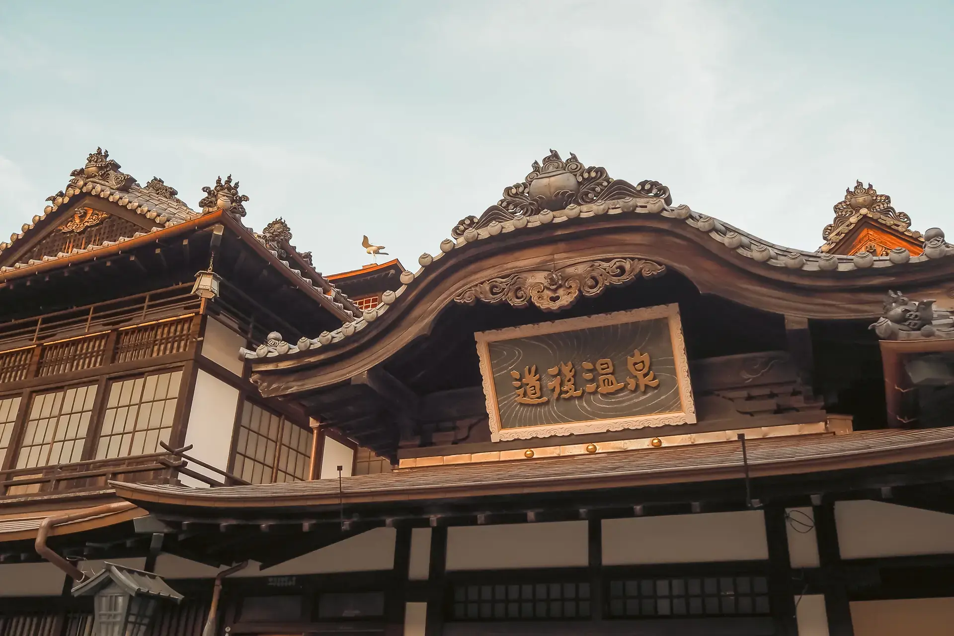 Traditional Japanese hot spring bathhouse featuring ornate curved rooflines with decorative tile eaves and a central wooden sign displaying Chinese characters, set against a tranquil evening sky.