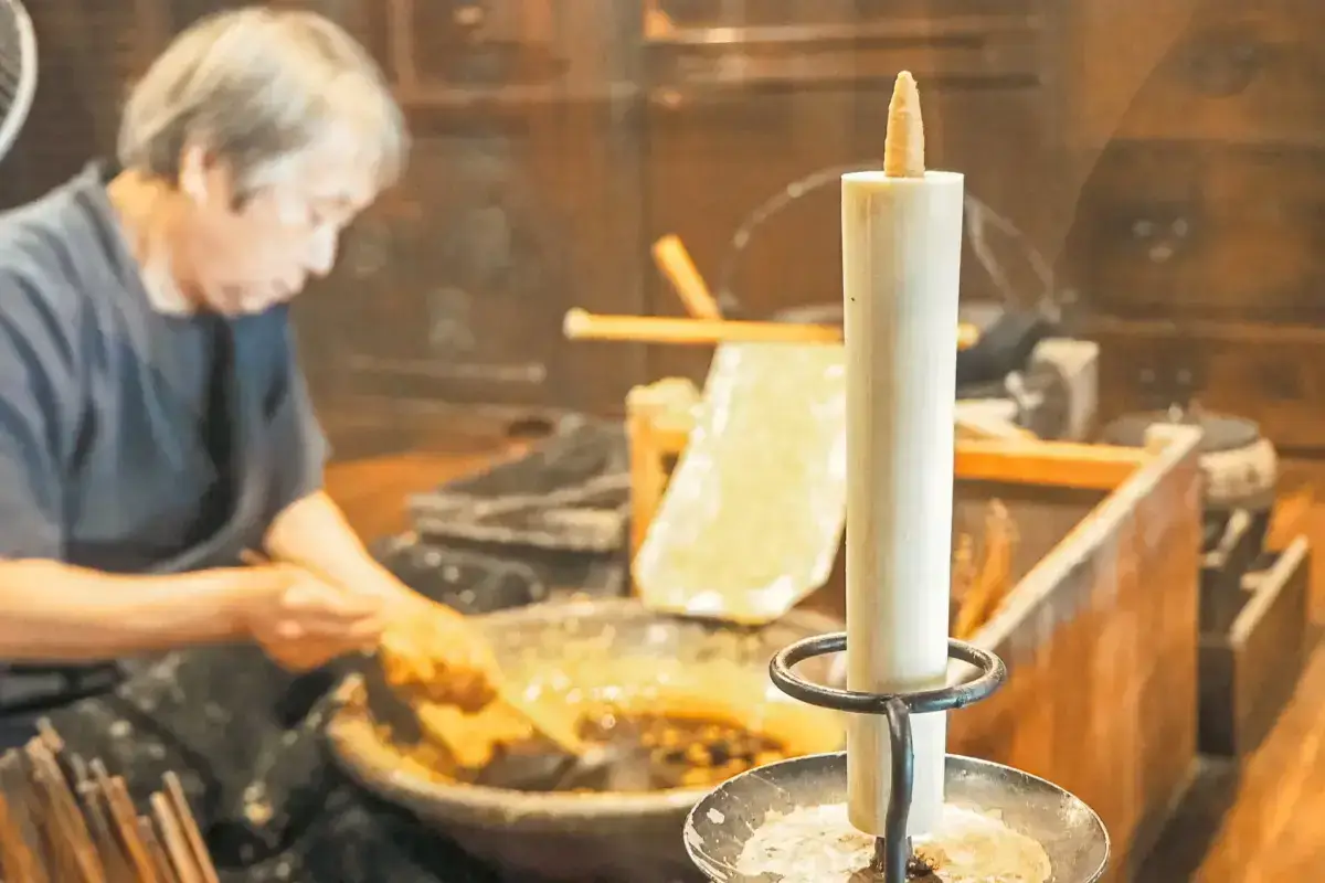 Elderly artisan craftsperson working at a traditional wooden workbench with tools and materials, with a lit white candle in an antique metal candleholder in the foreground creating a peaceful workshop atmosphere.