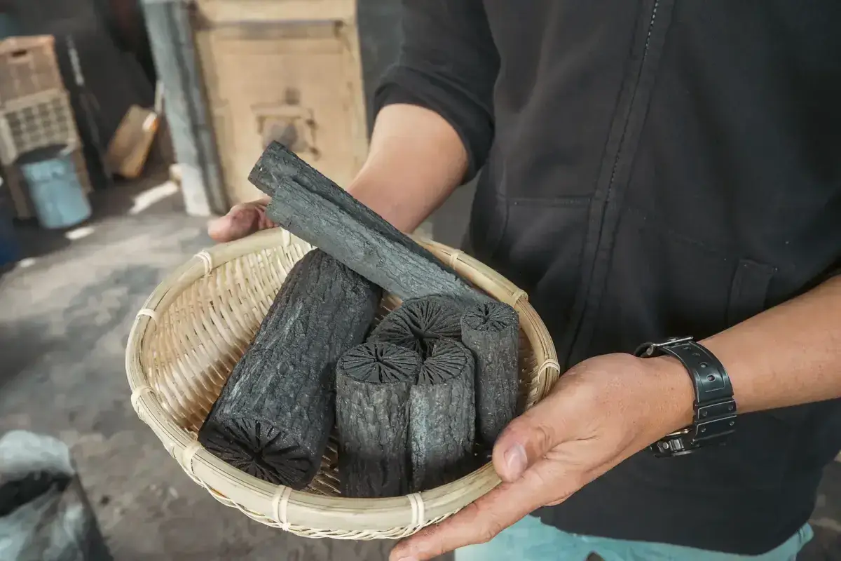 Hands holding a woven bamboo basket containing traditional Japanese binchotan charcoal pieces with distinctive radial patterns, displayed in a rustic workshop setting.