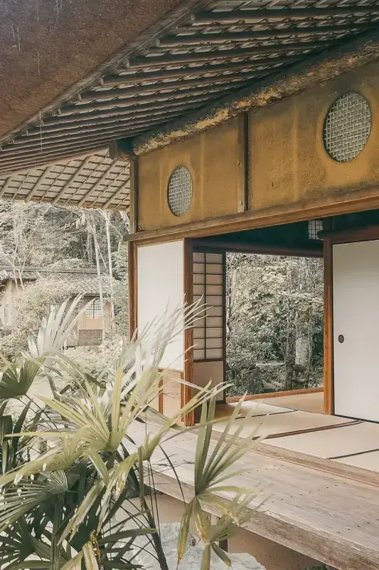 Traditional Japanese architecture with wooden beams and latticed windows, featuring a covered entrance with natural stone walls, wooden deck, and tropical plants in the foreground creating a serene garden atmosphere.