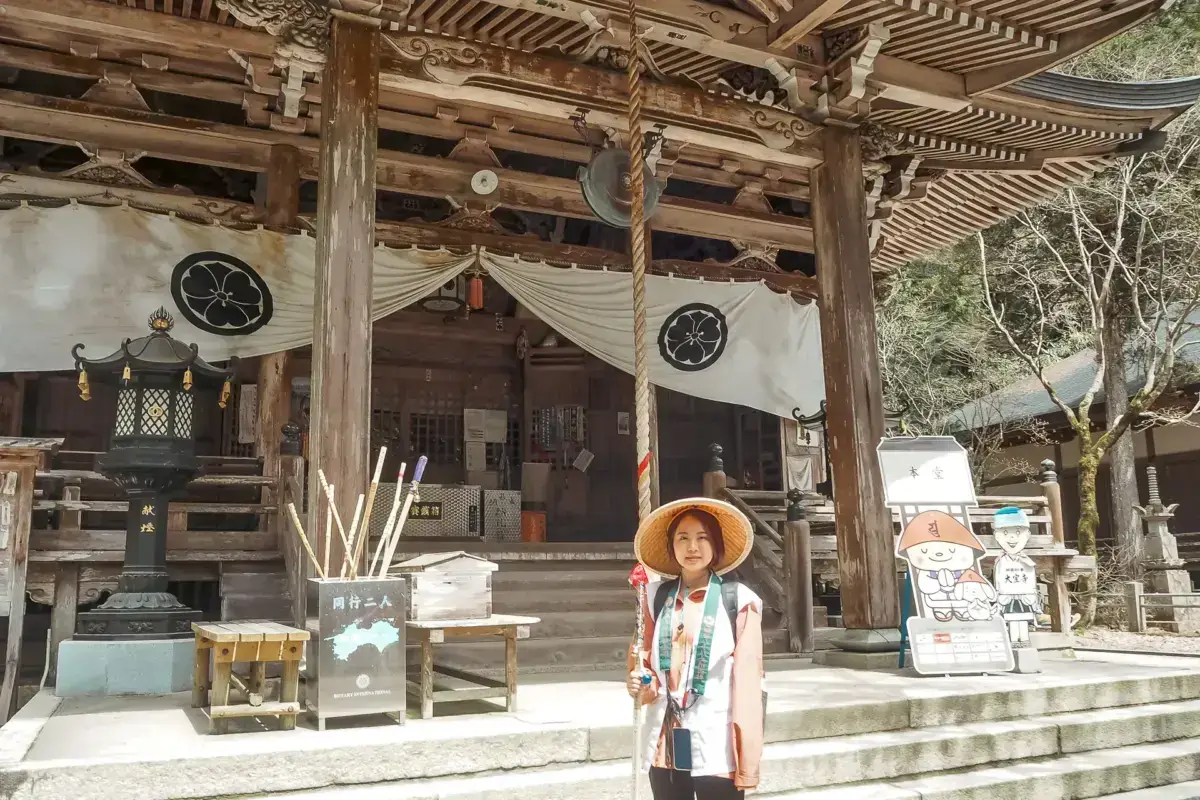 Female pilgrim wearing traditional straw hat and walking staff stands before a historic Japanese Buddhist temple with ornate wooden architecture and decorative curtains bearing clan crests.