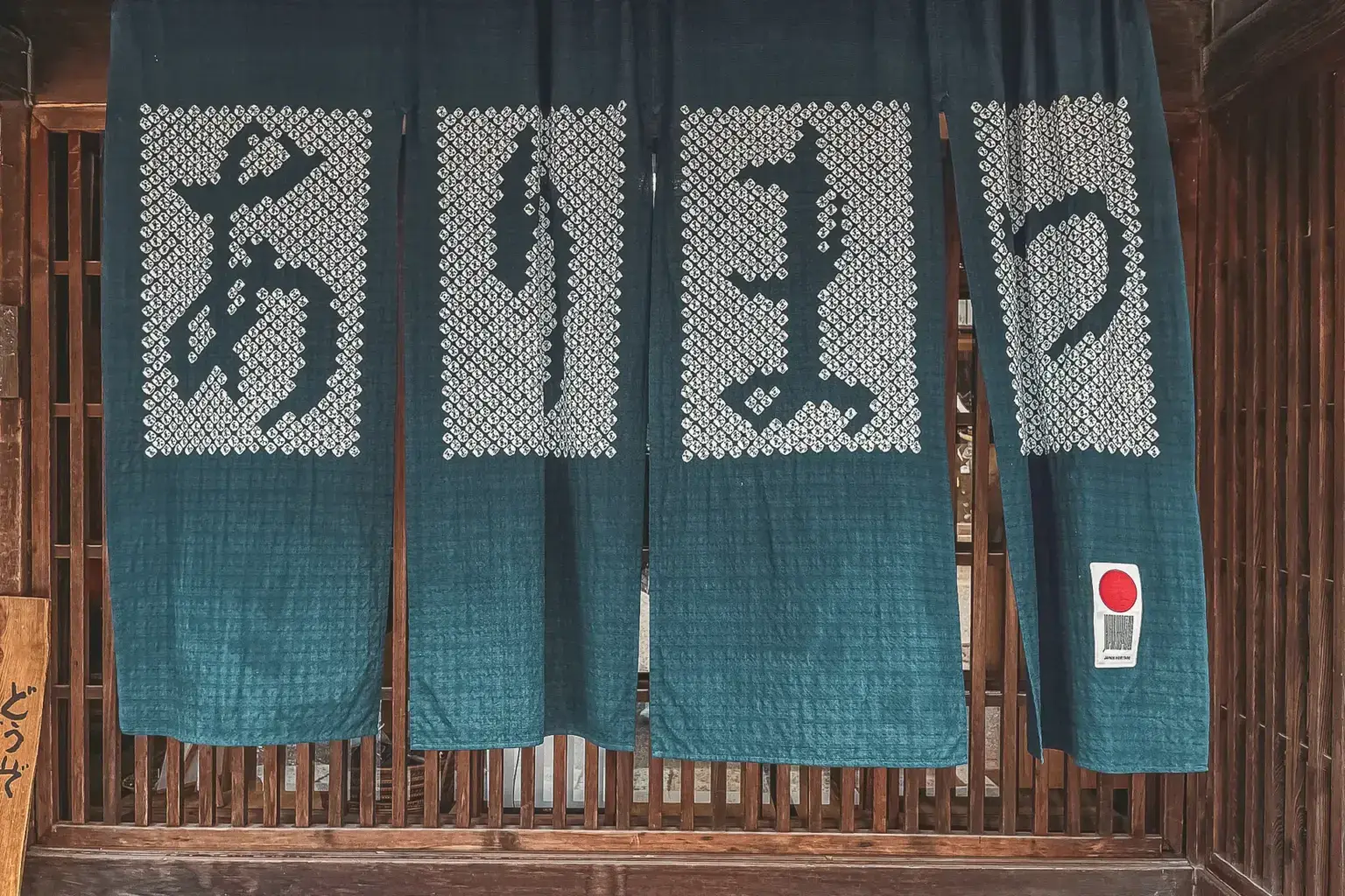 Traditional Japanese storefront entrance with four teal-blue noren curtains hanging from wooden slats, displaying white kanji characters, with a small red and white Japanese flag visible on the right panel.
