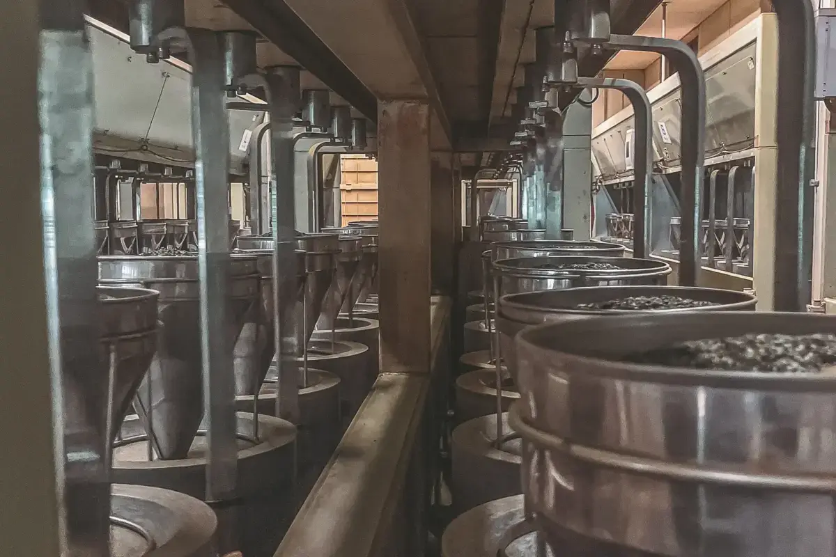 Traditional Japanese sake brewing facility with rows of metal fermentation tanks and industrial piping equipment in a wooden-beamed production room.