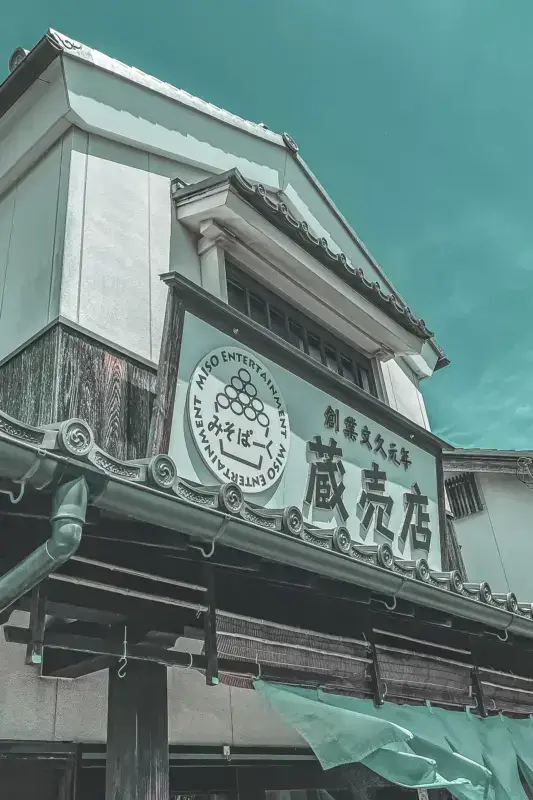 Traditional Japanese storefront in Aichi with distinctive architectural features including white walls, tiled roof with ornamental tiles, wooden lattice work, Japanese signage, and a circular logo displaying Miso Entertainment Group branding.