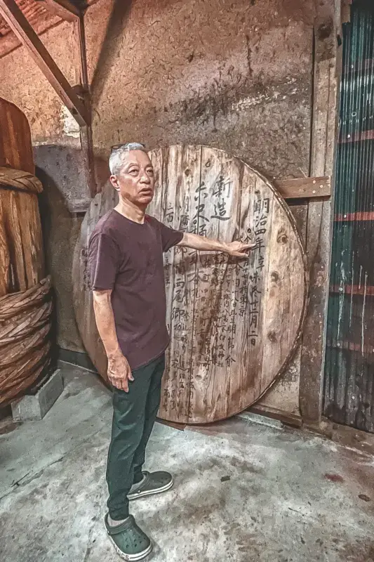 Elderly Japanese craftsman standing beside a large traditional wooden barrel with metal hoops in a rustic workshop with weathered walls and wooden beams.