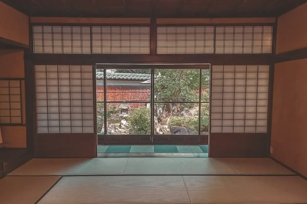 Traditional Japanese tatami room with shoji screens overlooking a serene garden with trees and a tiled-roof structure, exemplifying historic Aichi architecture.