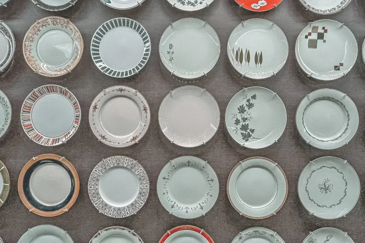 Overhead view of traditional Japanese ceramic plates arranged in neat rows, featuring various minimalist designs including delicate botanical patterns, geometric shapes, and subtle glazes in neutral tones.
