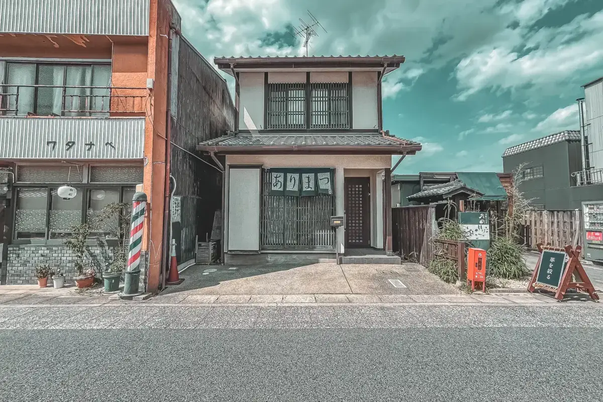 Traditional two-story Japanese storefront with distinctive latticed windows and signage, flanked by modern buildings under a dramatic cloudy sky.