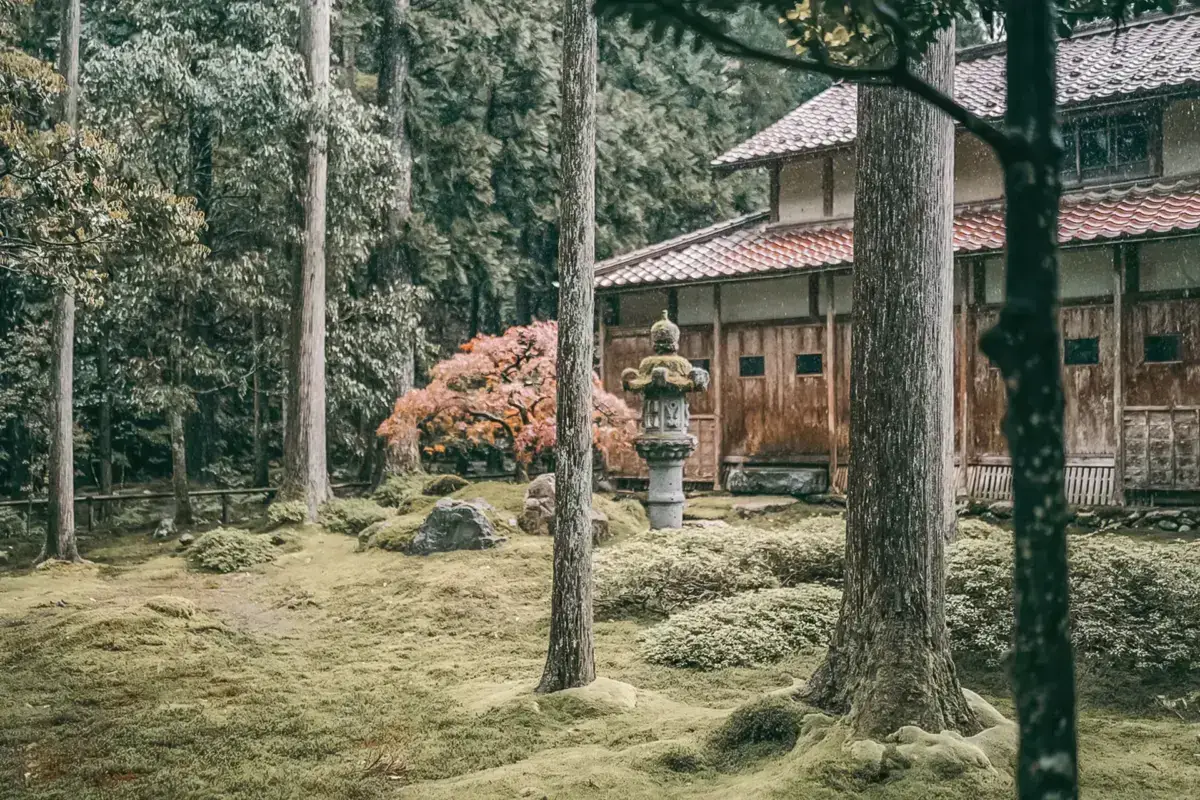 Traditional Japanese wooden temple or shrine building with distinctive tiled roof surrounded by tall trees and raked sand garden, featuring a stone lantern and vibrant autumn foliage.